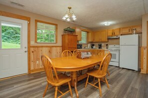 A dining area with a wooden table and chairs, a kitchen with wooden cabinets and appliances, and a window with blinds. - 🍃 Quaint Cabin on Cosby Creek 🏞️ Creekside Farm, Hot Tub & Park Access (Cosby)