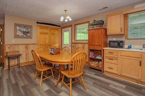 A dining area with a wooden table and chairs, a kitchen with wooden cabinets, a microwave, and a window with blinds. - 🍃 Quaint Cabin on Cosby Creek 🏞️ Creekside Farm, Hot Tub & Park Access (Cosby)