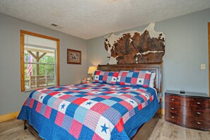 A bedroom with a bed, a window, a wall-mounted lamp, a wooden dresser, and a large piece of animal hide on the wall. - 🍃 Quaint Cabin on Cosby Creek 🏞️ Creekside Farm, Hot Tub & Park Access (Cosby)
