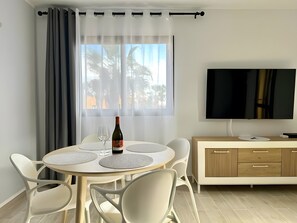 A modern living room with a round dining table, white chairs, a TV, and a cabinet. - B-rent Digital Boutique Casa Querida – Modern Comfort with Terrace & Pool (Corralejo)