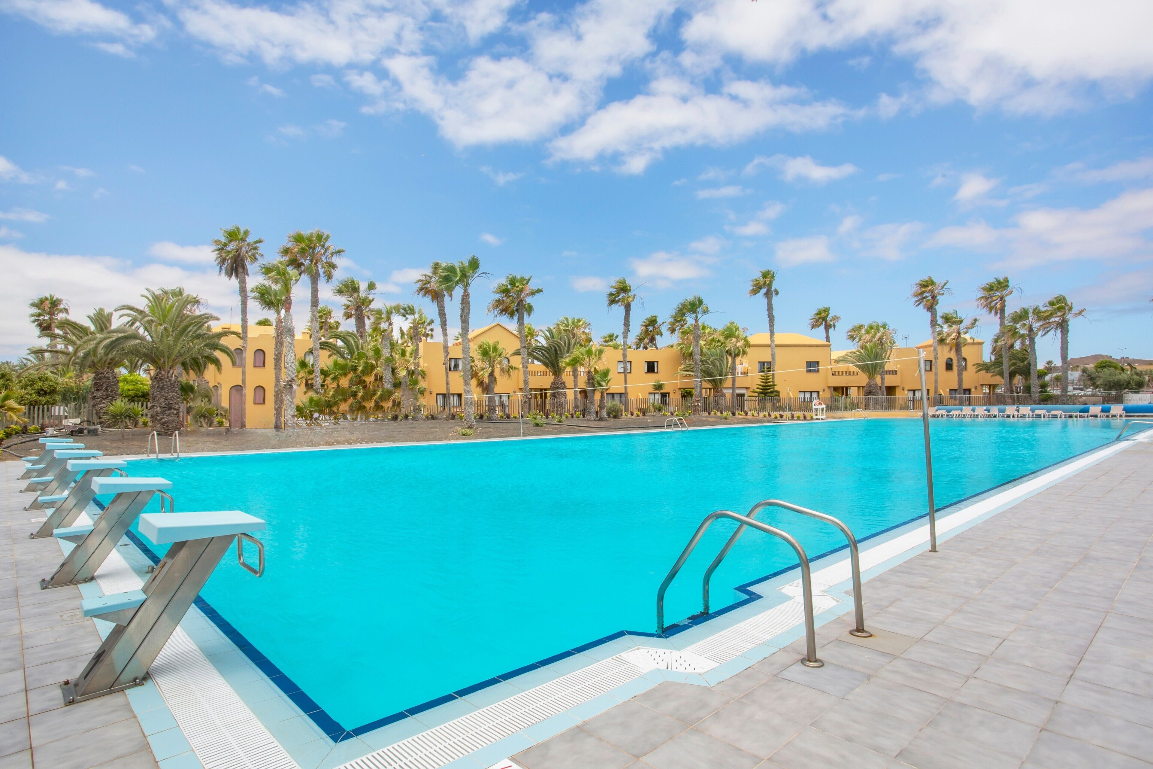 Ein Swimmingpool mit klarem, blauem Wasser, umgeben von Palmen und gelben Gebäuden.