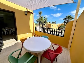 A balcony with a white table and chairs, a view of palm trees, and a yellow building. - B-rent Digital Boutique Casa Querida – Modern Comfort with Terrace & Pool (Corralejo)