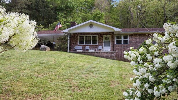 A brick house with a covered porch, a flag, and white flowering bushes. - 🌲 70 Acre Mountain Farmstead 🏞️ Stocked Pond, Game Room & Seclusion (Sevierville)