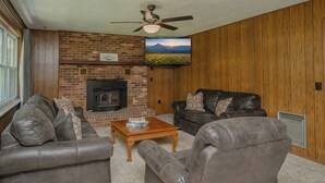 A living room with a fireplace, leather sofas, a coffee table, and a television mounted on the wall. - 🌲 70 Acre Mountain Farmstead 🏞️ Stocked Pond, Game Room & Seclusion (Sevierville)