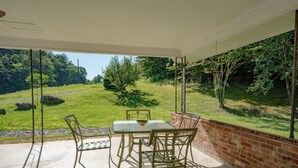 A covered patio with a table and chairs, overlooking a grassy landscape with trees and a clear sky. - 🌲 70 Acre Mountain Farmstead 🏞️ Stocked Pond, Game Room & Seclusion (Sevierville)