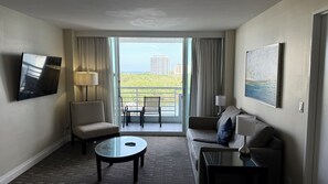Een hotelkamer met een bank, fauteuil, salontafel, televisie en een balkon met uitzicht op gebouwen en groen.