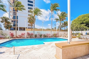 Una piscina con agua azul cristalina, rodeada de palmeras y un edificio blanco con balcones.