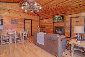 Ein Blockhäuschen-Interieur mit Holzbalkendecke, Kamin, einer Couch, einem Esstisch mit Stühlen und einem an der Wand befestigten Fernseher.