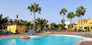 A pool area with lounge chairs, palm trees, and yellow buildings. - B-Rent El Royal Corralejo: Comfort and Style in a 1-Bedroom Apartment (Corralejo)