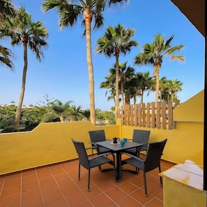 A patio with a table and chairs, surrounded by palm trees and a yellow wall. - B-Rent El Royal Corralejo: Comfort and Style in a 1-Bedroom Apartment (Corralejo)