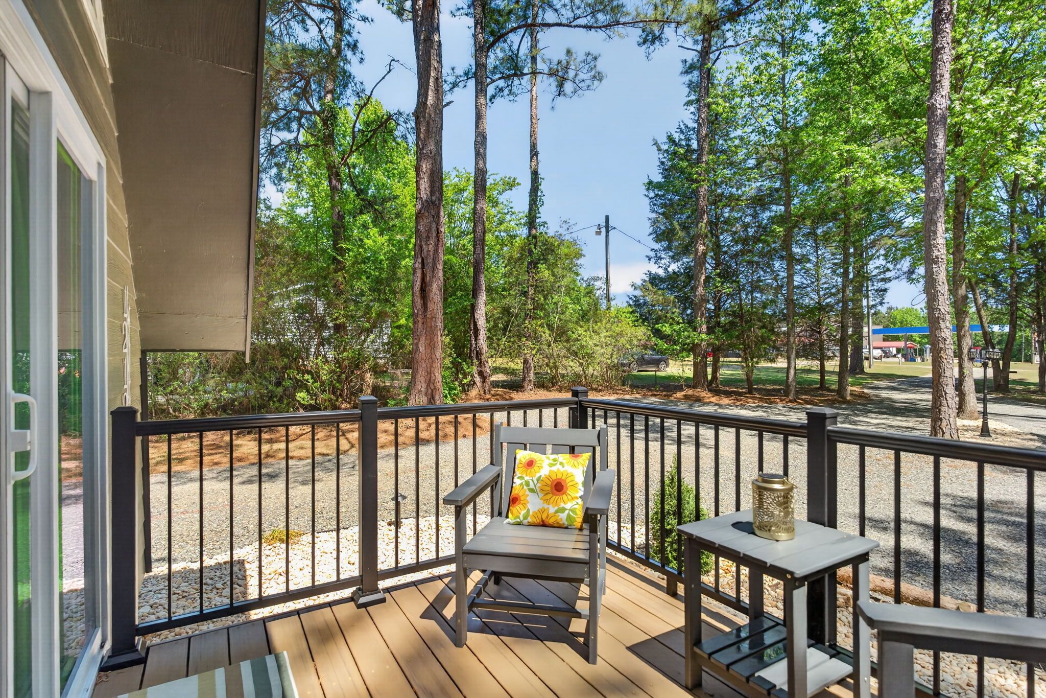 A wooden deck with a chair and table, surrounded by a black railing, overlooking a gravel path and trees.