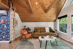 Ein Loft-Zimmer mit Holzbalkendecke, einem braunen Ledersofa, einem weißen Couchtisch und einem Fenster mit Blick auf Grünflächen.