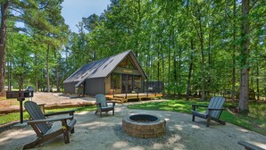 A cabin with a deck, surrounded by trees, featuring Adirondack chairs and a fire pit.