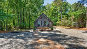 A cabin with a triangular roof, a porch with railings, and a gravel driveway surrounded by trees.