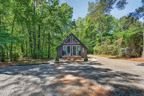 A cabin with a triangular roof, a porch with railings, and a gravel driveway surrounded by trees.