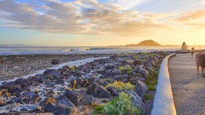 A rocky beach with a paved walkway, a distant mountain, and the ocean. - B-Rent Bristol Beach Surf & Fun – Your Perfect Fuerteventura Getaway (Corralejo)