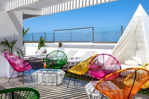 Terraço no telhado com cadeiras coloridas, um toldo branco e vista para o mar.