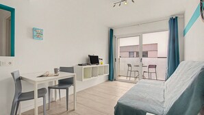 A room with a bed, a desk, two chairs, a computer, and a view through a glass door to an outdoor area with a table and chairs. - B-Rent Bristol Beach Surf & Fun – Your Perfect Fuerteventura Getaway (Corralejo)
