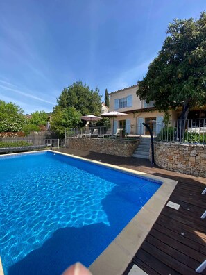 A swimming pool with a wooden deck, a stone wall, and a building with blue shutters. - Cozy family villa in Provence with a large terrace, pool, and village life right nearby. (Cotignac)