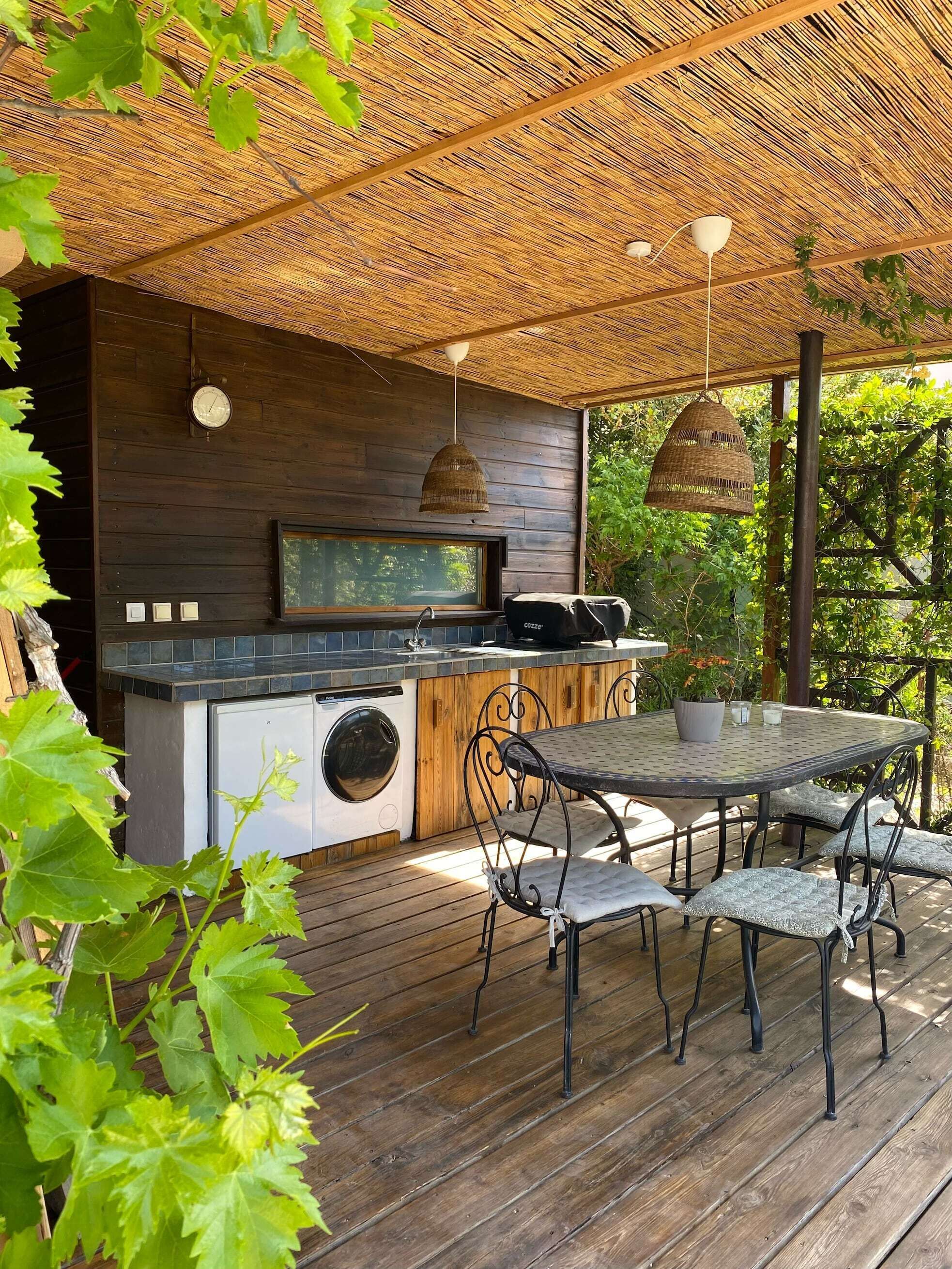 An outdoor kitchen with a built-in washing machine, a dining table with chairs, and hanging pendant lights.