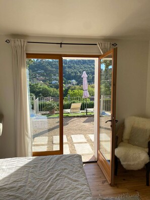 A bedroom with a bed, a chair, and a sliding glass door leading to a balcony with a view of a hillside and a garden. - Cozy family villa in Provence with a large terrace, pool, and village life right nearby. (Cotignac)