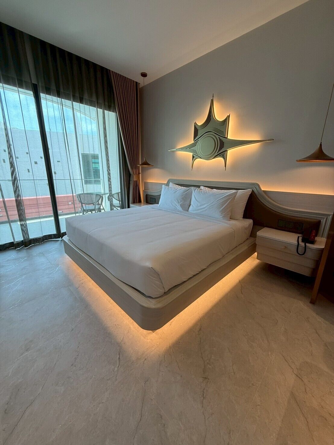 A modern hotel room with a large bed, a nightstand, a wall-mounted star light, and a view of the outside through large windows.