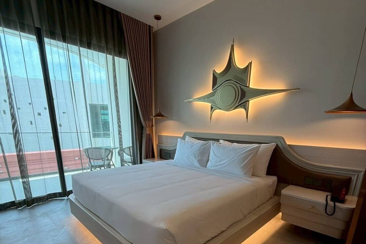 A modern hotel room with a large bed, a nightstand, a wall-mounted star light, and a view of the outside through large windows.