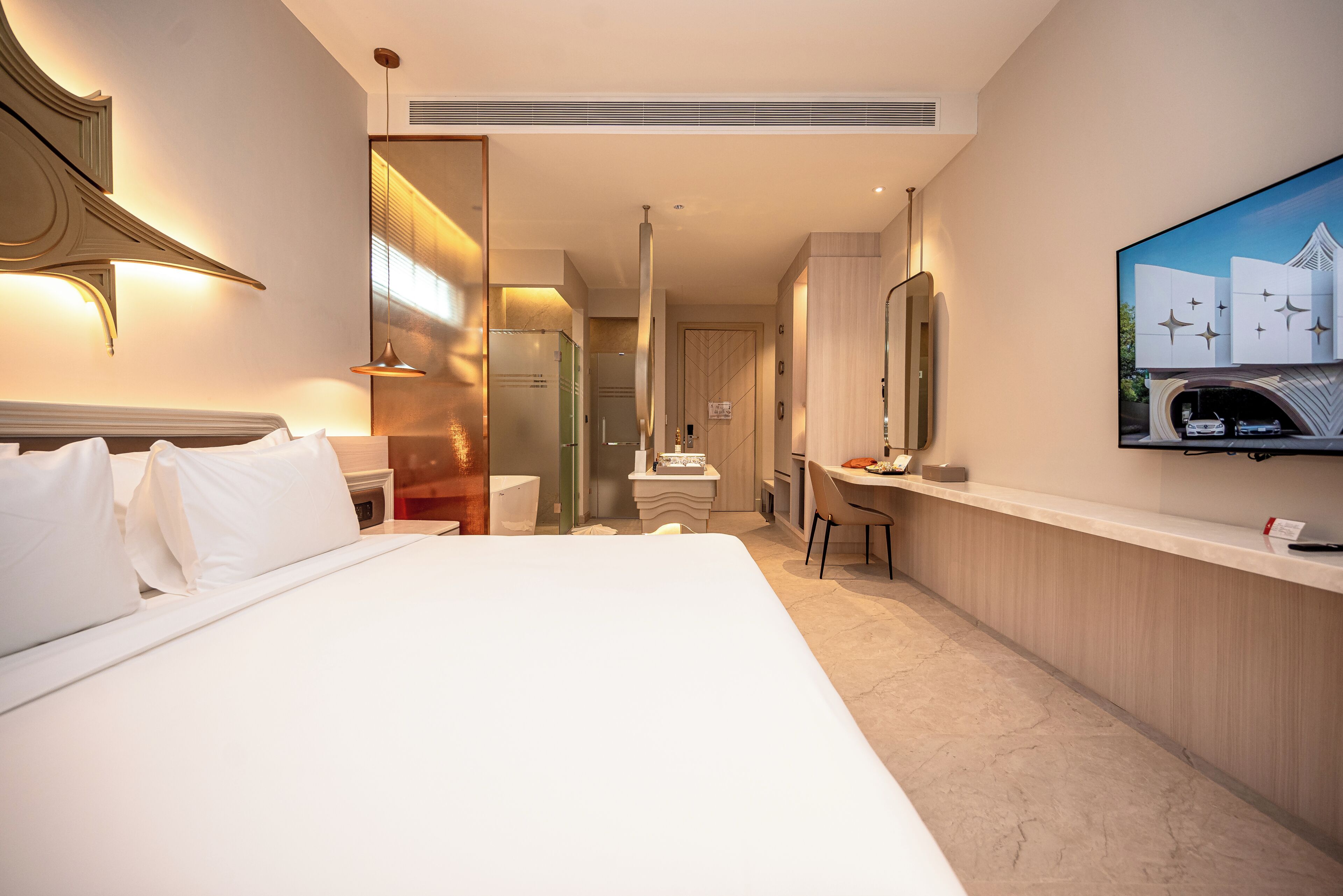 A hotel room with a large bed, a desk, a chair, a television, and a bathroom visible through a glass partition.