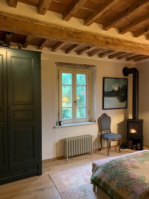 A room with a wooden ceiling, a large window, a radiator, a chair, a bed with a patterned blanket, and a fireplace. - Quercus Suite - Le Gusciane Country Inn (Toscana)