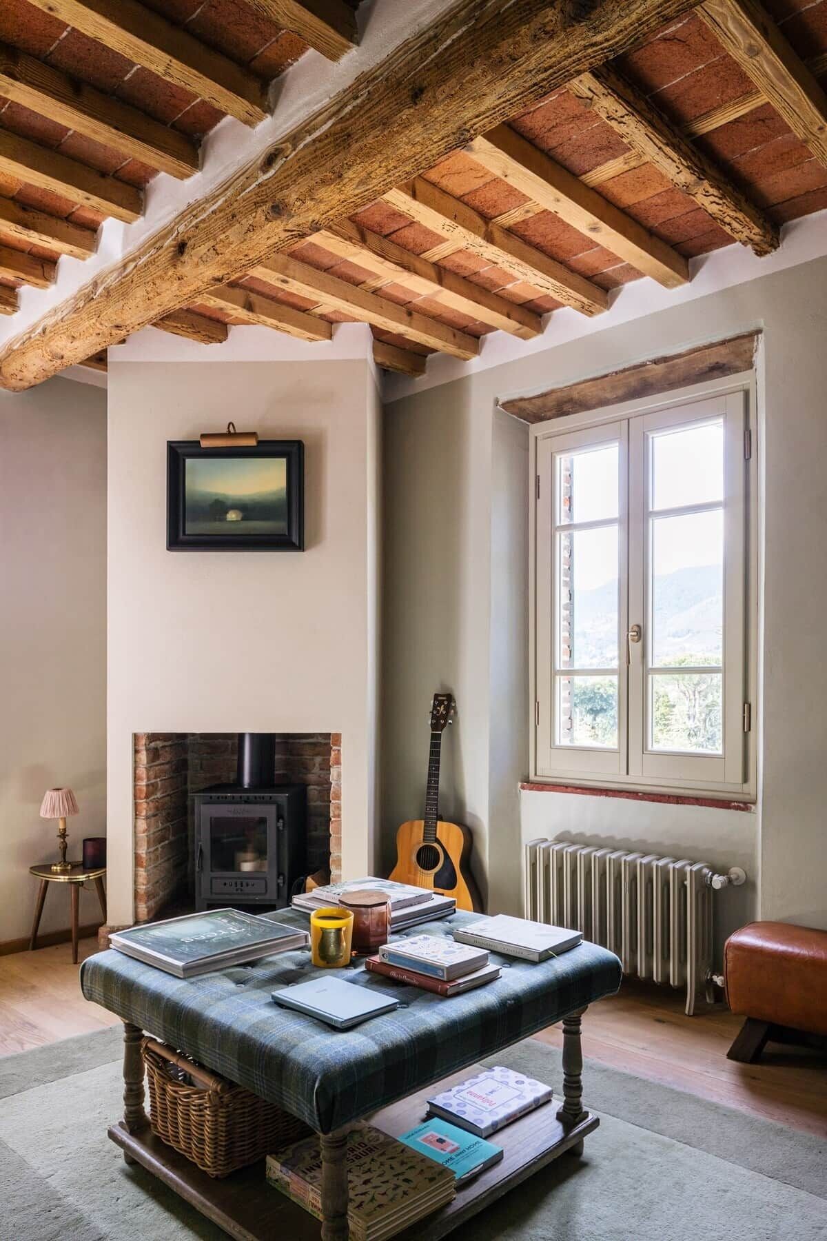 Une pièce chaleureuse avec une cheminée, une guitare, une table remplie de livres et une fenêtre donnant sur un beau paysage.