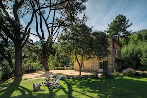 Una casa de pedra rústica amb un jardí, un pati amb taula i cadires, i un arbre gran al primer pla.