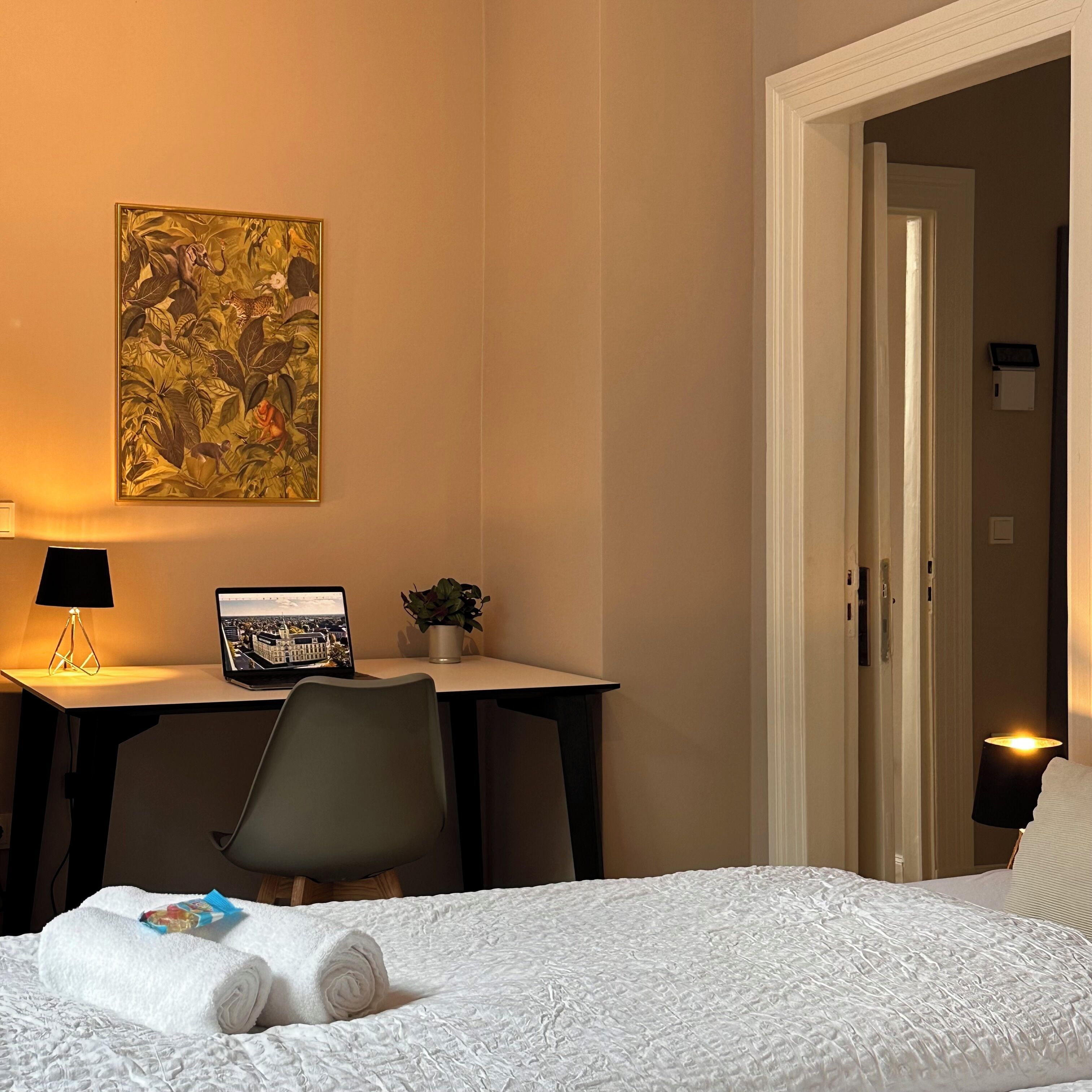 Una habitación de hotel con una cama, un escritorio con una computadora portátil, una silla, un cuadro en la pared y una puerta que da a otra habitación.