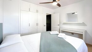 Une chambre à coucher avec un lit, une armoire blanche, un miroir au-dessus de la commode et un ventilateur de plafond.