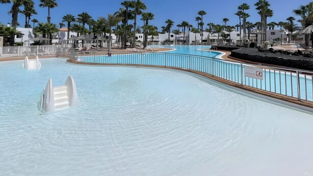 Une grande piscine incurvée, dotée d’une terrasse blanche et de rampes en métal, entourée de palmiers et de bâtiments blancs.