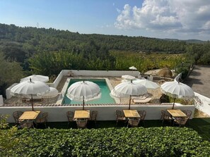 A rooftop pool with white umbrellas and wicker chairs, surrounded by lush greenery and a hilly landscape.