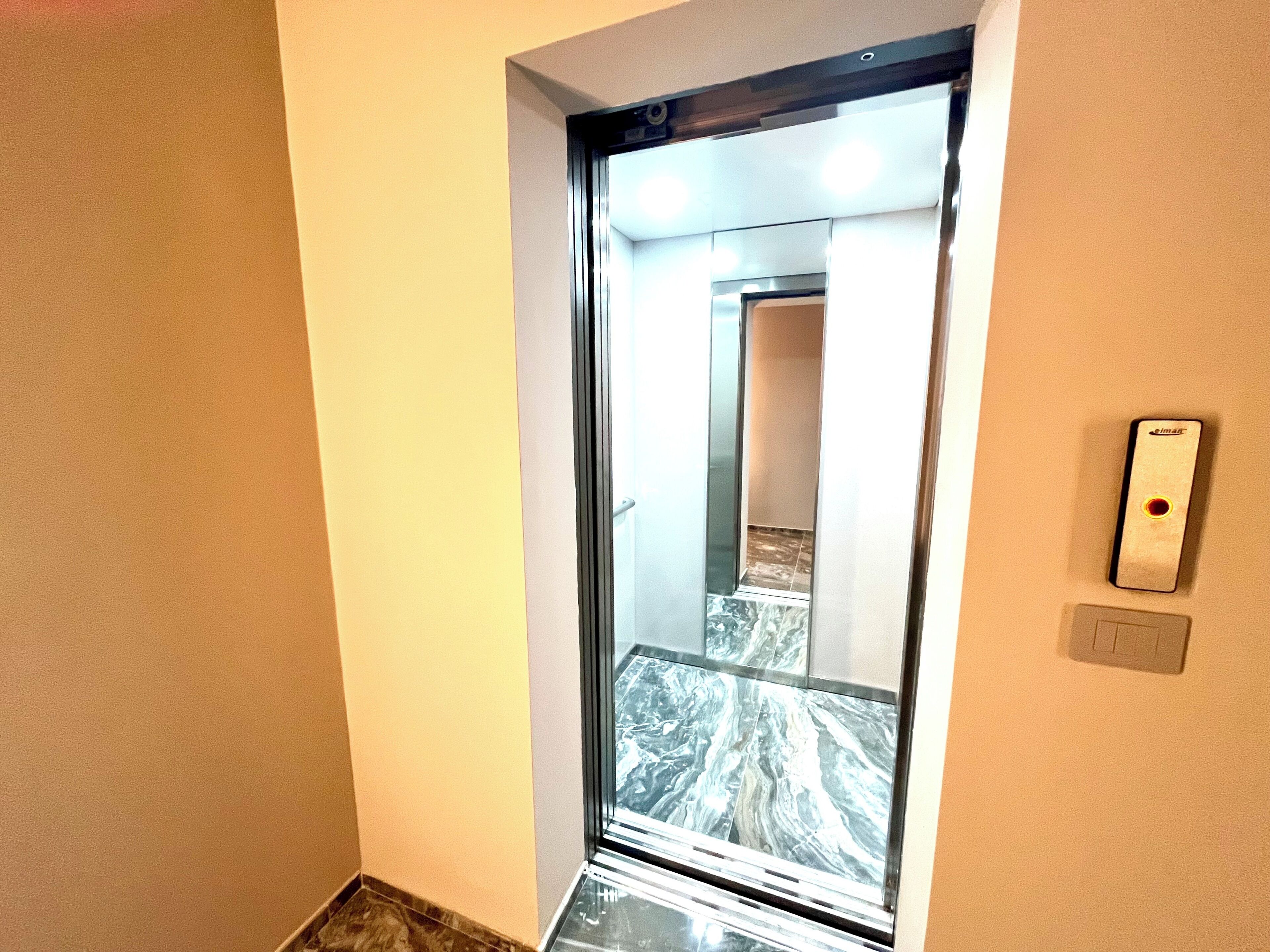 Un corridoio stretto con una porta a vetro per l'ascensore, un pavimento in marmo e un pulsante dell'ascensore fissato al muro.