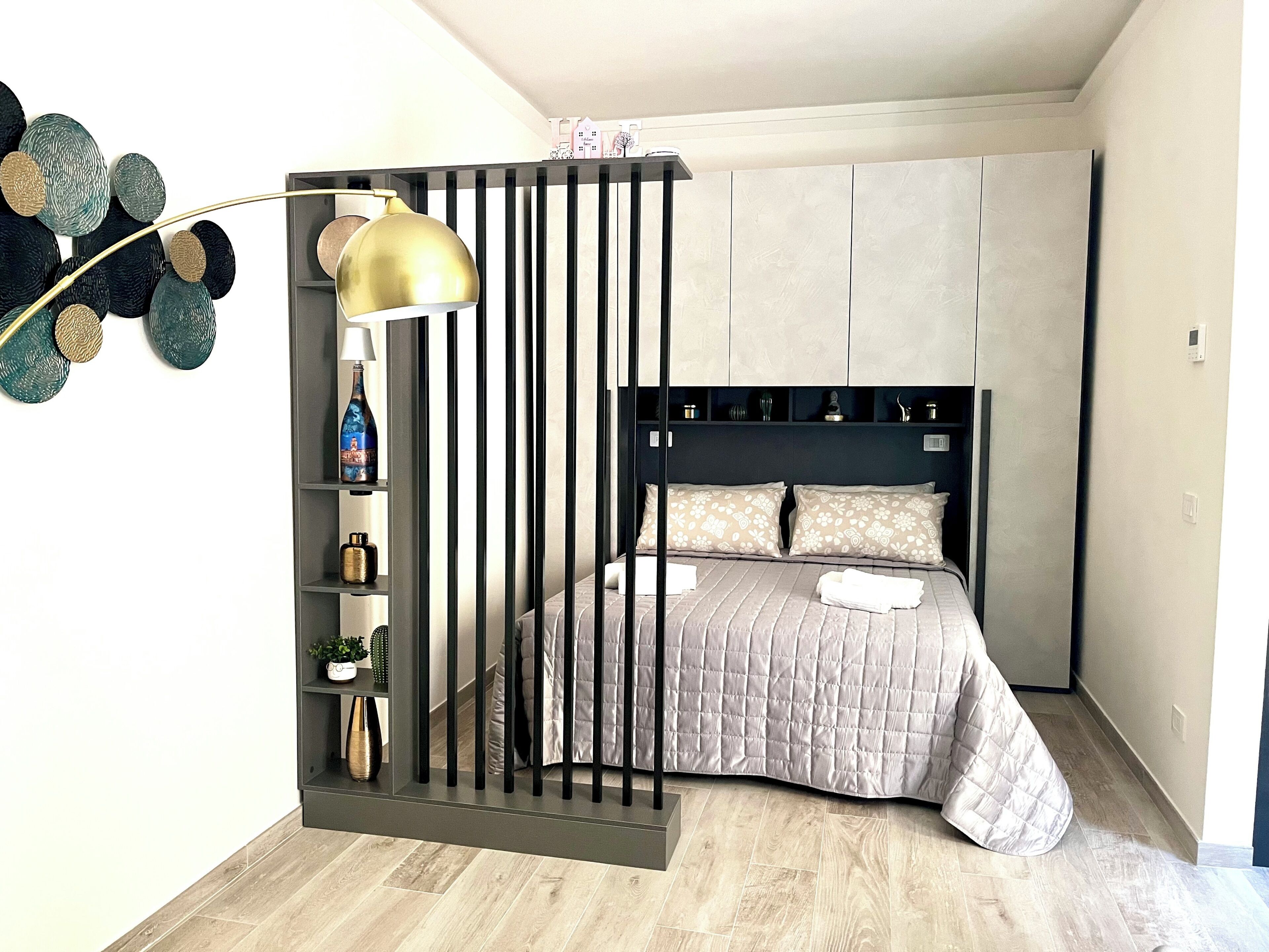 A bedroom with a bed, a bedside table, a lamp, and a wall-mounted shelf with decorative items.