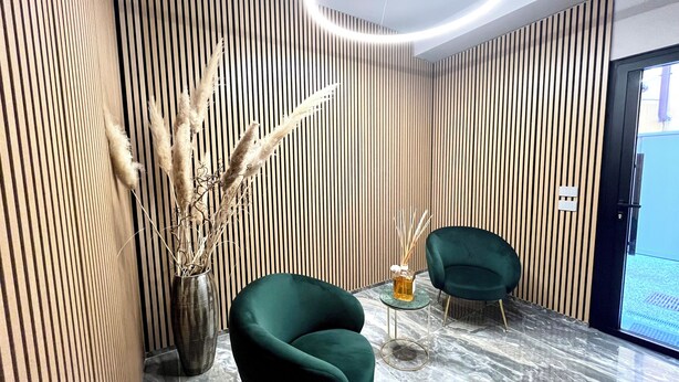 A modern interior with vertical wooden paneling, a marble floor, two green armchairs, a vase with dried plants, and a glass table. - Garibaldi Living - Centro Storico Parma (Parma)