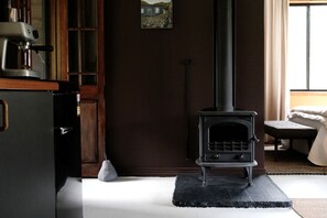 Una habitación con estufa de leña, una mesita, una silla y un ventanal.