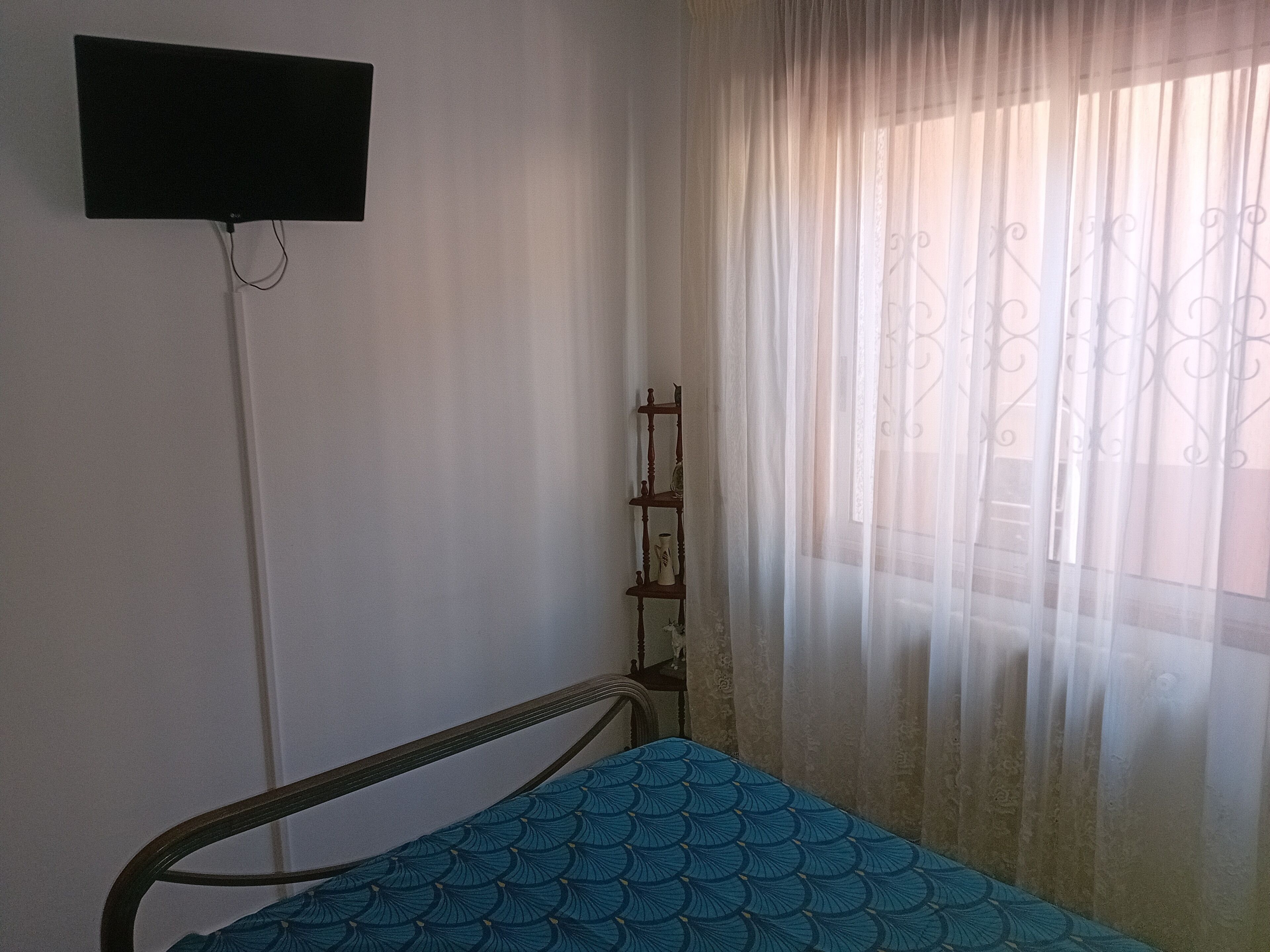 Ein Einzelbett mit einem gemusterten Überwurf, ein Nachttisch, ein an der Wand montierter Fernseher und durchscheinende Vorhänge mit einem dekorativen Fensterrollo.