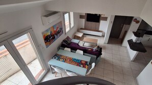 Et rum med højt til loftet, et spisebord, stole, en sofa, et tv ophængt på væggen og et køkkenområde.