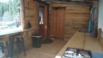 Una cabaña rústica con interior de madera, una zona de comedor con taburetes, una estufa y una puerta que da al exterior.