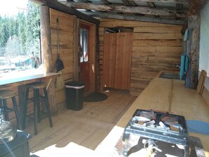 Una cabana rústica amb interior de fusta, una zona de menjador amb tamborets, una cuina i una porta que dóna a l'exterior.