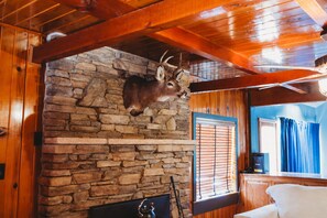 A stone fireplace with a mounted deer head, wooden beams, and a window with blinds. - Bigfoot Alley | Movie Room + Walkable Location (Shaver Lake)