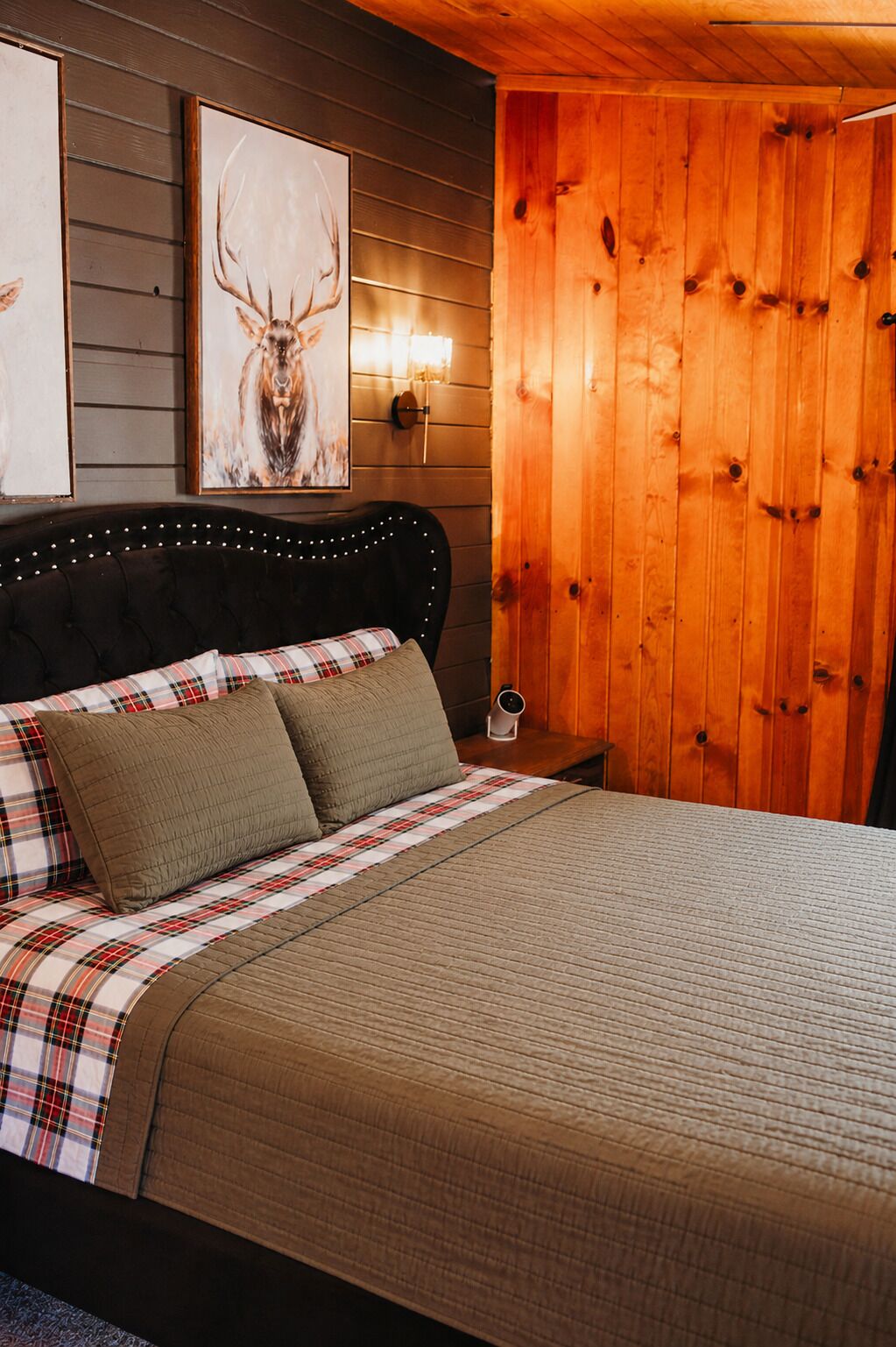 A bed with a plaid bedspread, two pillows, a headboard with lights, a wooden wall panel, and a framed deer picture.