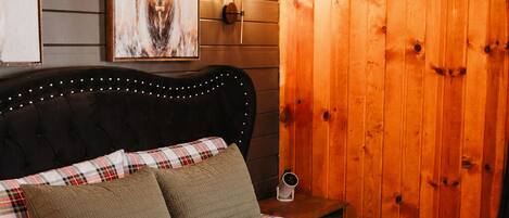 A bed with a plaid bedspread, two pillows, a headboard with lights, a wooden wall panel, and a framed deer picture.