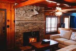 A cozy cabin interior with a stone fireplace, wooden walls, a ceiling fan, and a sofa. - Bigfoot Alley | Movie Room + Walkable Location (Shaver Lake)