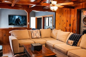 A cozy living room with a sectional sofa, a wooden coffee table, and a television mounted on the wall. - Bigfoot Alley | Movie Room + Walkable Location (Shaver Lake)
