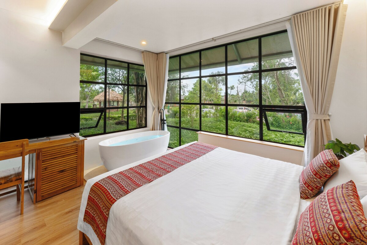Un dormitorio con una cama grande, una bañera, un televisor y vistas a la vegetación.