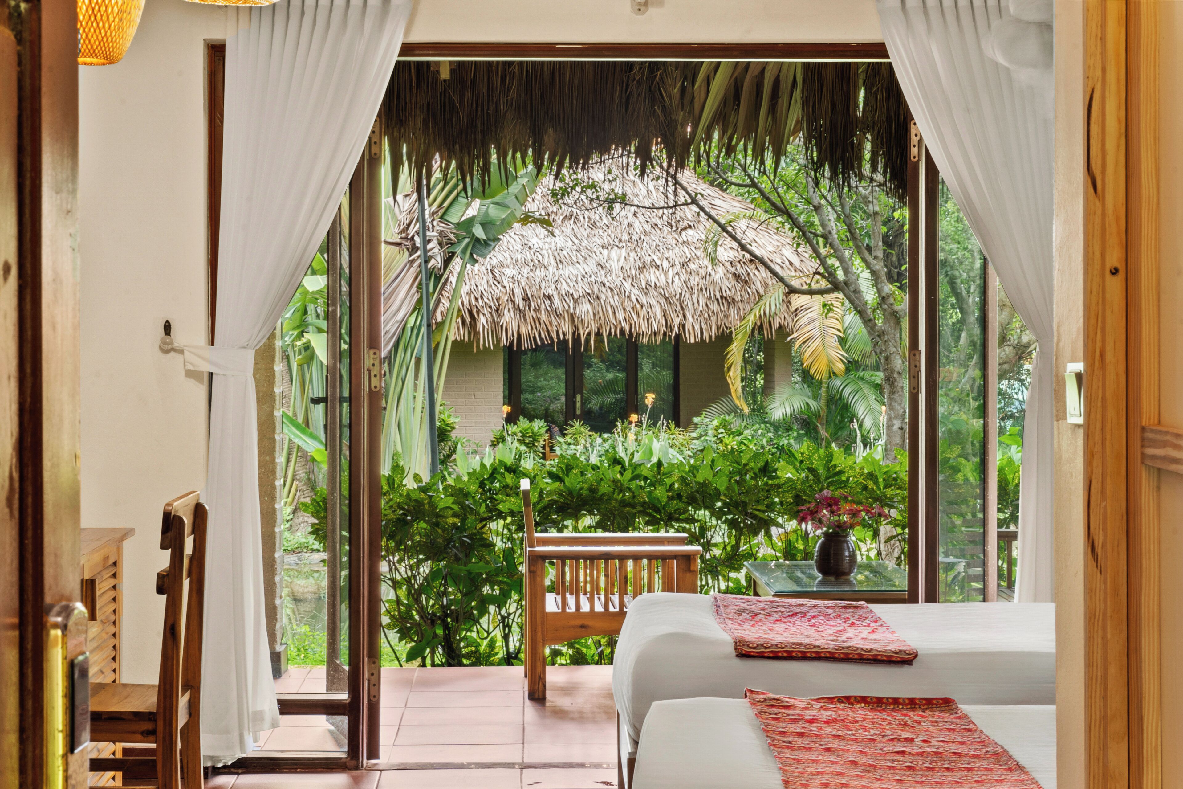 A room with a bed, a wooden chair, a table, and a view of lush greenery and a thatched roof structure.